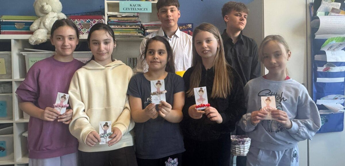 Uczniowie stoją w szkolnej sali przy kąciku czytelniczym, prezentując własnoręcznie przygotowane upominki z okazji Dnia Kobiet.