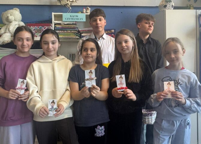 Uczniowie stoją w szkolnej sali przy kąciku czytelniczym, prezentując własnoręcznie przygotowane upominki z okazji Dnia Kobiet.