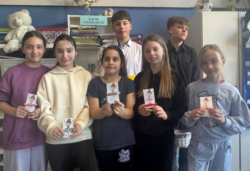 Uczniowie stoją w szkolnej sali przy kąciku czytelniczym, prezentując własnoręcznie przygotowane upominki z okazji Dnia Kobiet.