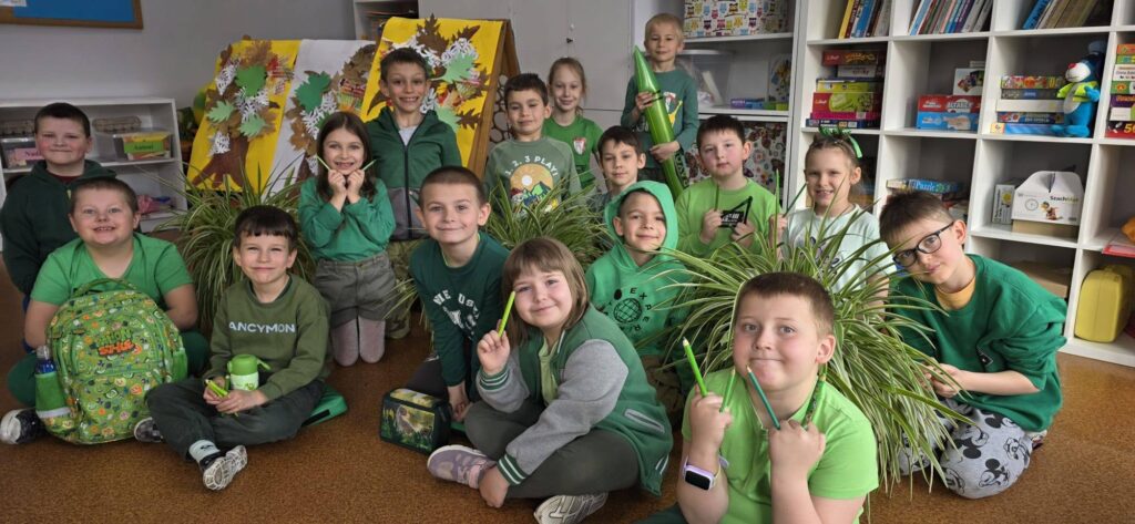 Uczniowie klasy młodszej w sali lekcyjnej, ubrani na zielono, pozują do zdjęcia wśród roślin i prac plastycznych związanych z tematyką wiosenną.