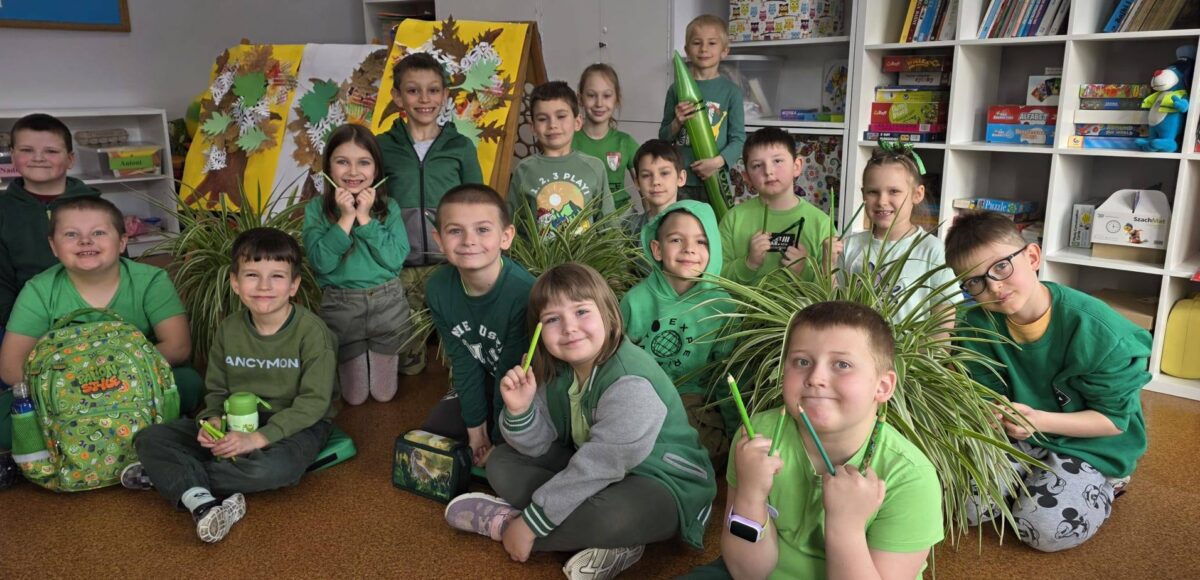 Uczniowie klasy młodszej w sali lekcyjnej, ubrani na zielono, pozują do zdjęcia wśród roślin i prac plastycznych związanych z tematyką wiosenną.