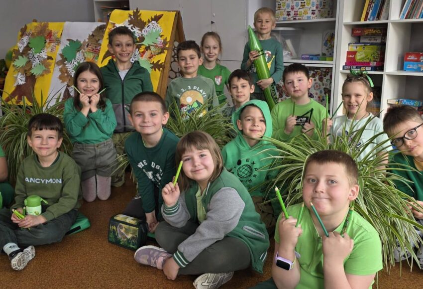 Uczniowie klasy młodszej w sali lekcyjnej, ubrani na zielono, pozują do zdjęcia wśród roślin i prac plastycznych związanych z tematyką wiosenną.