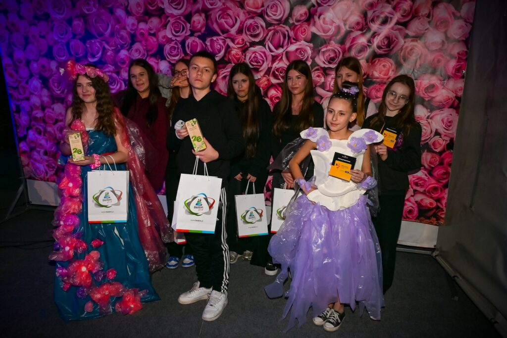 Uczniowie prezentują ekologiczne stroje wykonane z materiałów z recyklingu podczas konkursu Recycled Fashion Day, trzymając dyplomy i nagrody.