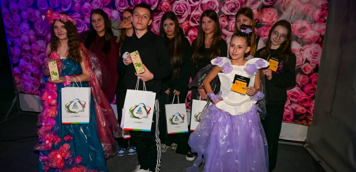 Uczniowie prezentują ekologiczne stroje wykonane z materiałów z recyklingu podczas konkursu Recycled Fashion Day, trzymając dyplomy i nagrody.