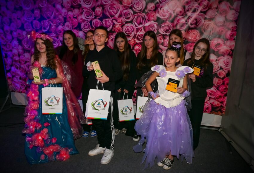 Uczniowie prezentują ekologiczne stroje wykonane z materiałów z recyklingu podczas konkursu Recycled Fashion Day, trzymając dyplomy i nagrody.