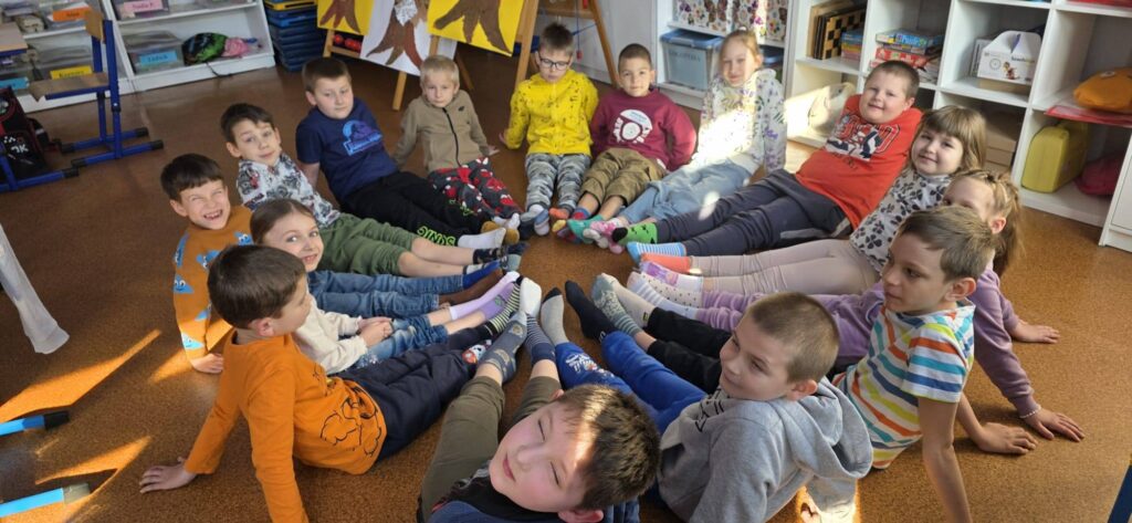 Uczniowie klasy młodszej siedzą w kręgu na podłodze w sali lekcyjnej, prezentując kolorowe, niepasujące do siebie skarpetki w ramach Dnia Skarpetek Nie do Pary.