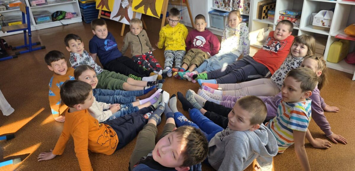 Uczniowie klasy młodszej siedzą w kręgu na podłodze w sali lekcyjnej, prezentując kolorowe, niepasujące do siebie skarpetki w ramach Dnia Skarpetek Nie do Pary.