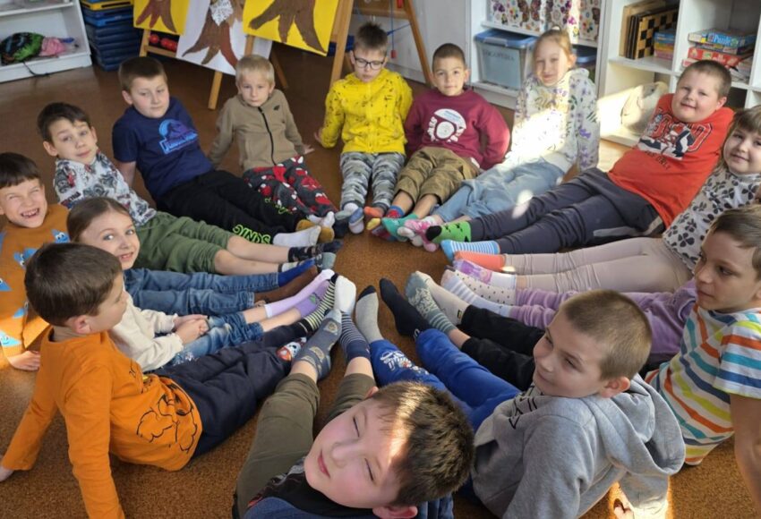 Uczniowie klasy młodszej siedzą w kręgu na podłodze w sali lekcyjnej, prezentując kolorowe, niepasujące do siebie skarpetki w ramach Dnia Skarpetek Nie do Pary.