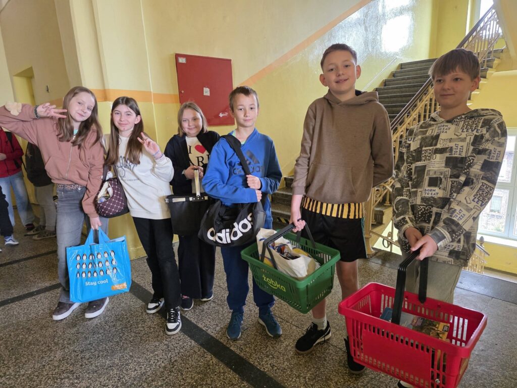 Uczniowie stoją na szkolnym korytarzu, trzymając książki i przybory w nietypowych przedmiotach, takich jak koszyki zakupowe, torby i walizki, w ramach Dnia bez plecaka.