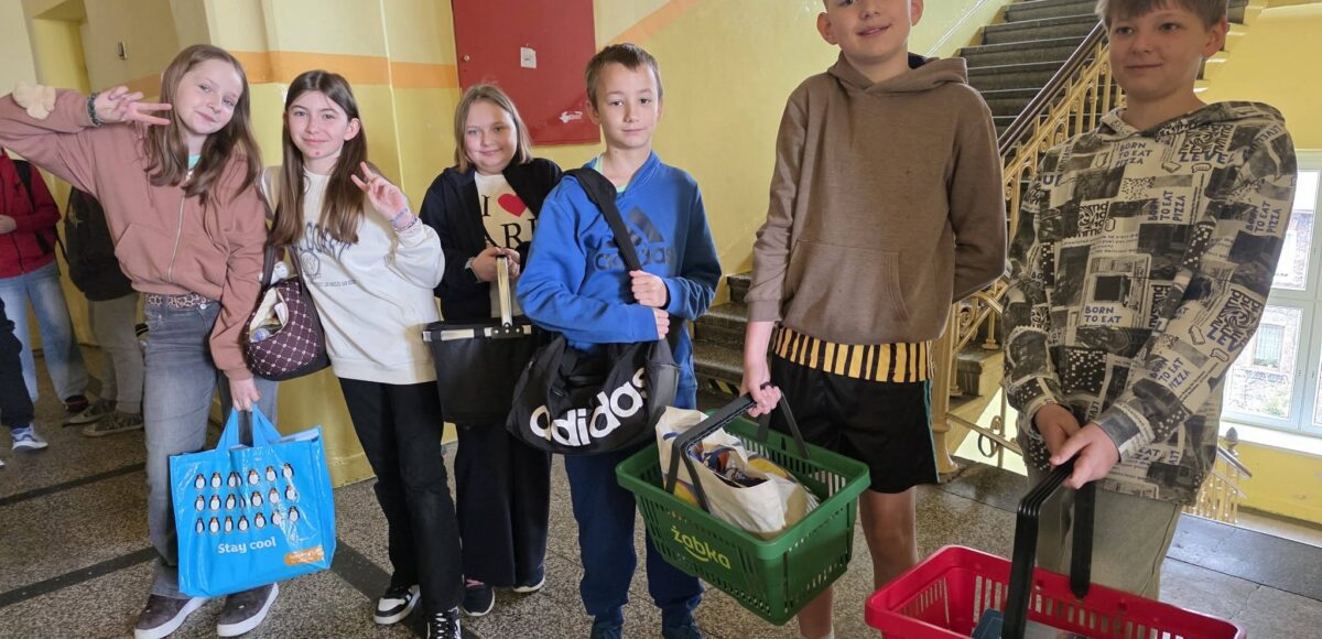 Uczniowie stoją na szkolnym korytarzu, trzymając książki i przybory w nietypowych przedmiotach, takich jak koszyki zakupowe, torby i walizki, w ramach Dnia bez plecaka.