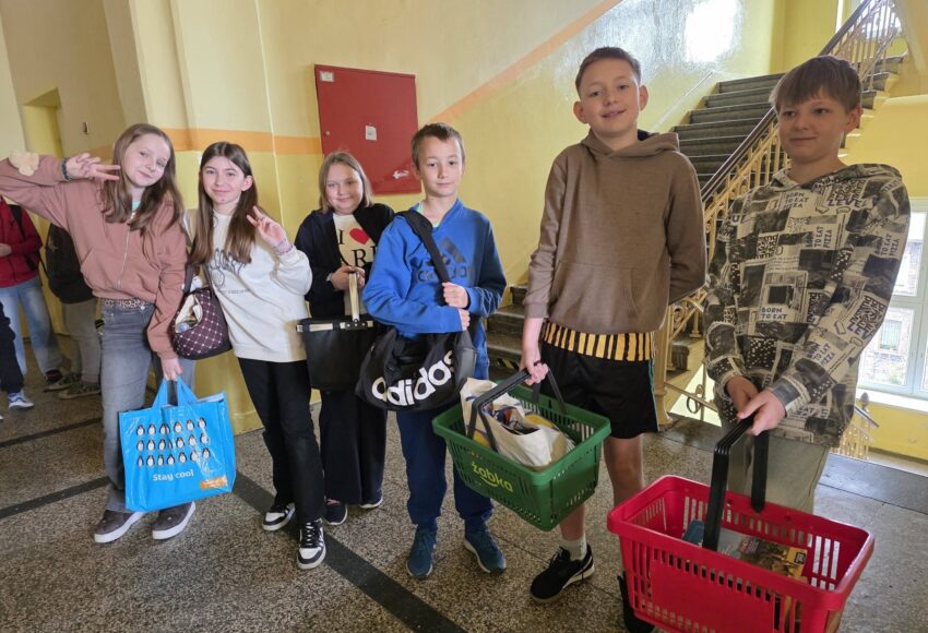 Uczniowie stoją na szkolnym korytarzu, trzymając książki i przybory w nietypowych przedmiotach, takich jak koszyki zakupowe, torby i walizki, w ramach Dnia bez plecaka.