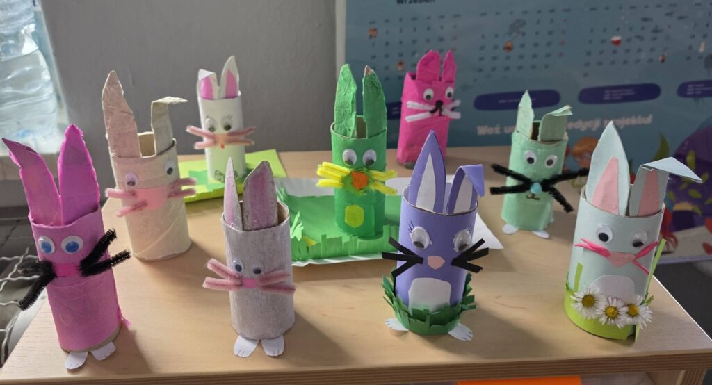 Kolorowe wielkanocne zajączki wykonane z rolek po papierze przez uczniów klasy 2, ozdobione papierem, ruchomymi oczkami i elementami dekoracyjnymi, ustawione na ławce szkolnej.