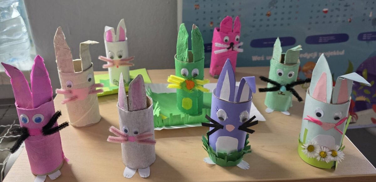 Kolorowe wielkanocne zajączki wykonane z rolek po papierze przez uczniów klasy 2, ozdobione papierem, ruchomymi oczkami i elementami dekoracyjnymi, ustawione na ławce szkolnej.