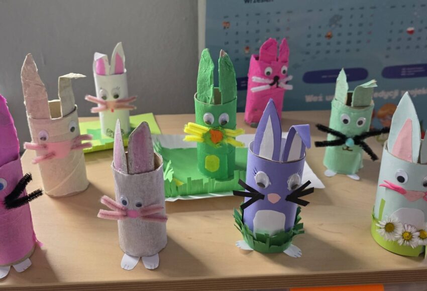Kolorowe wielkanocne zajączki wykonane z rolek po papierze przez uczniów klasy 2, ozdobione papierem, ruchomymi oczkami i elementami dekoracyjnymi, ustawione na ławce szkolnej.