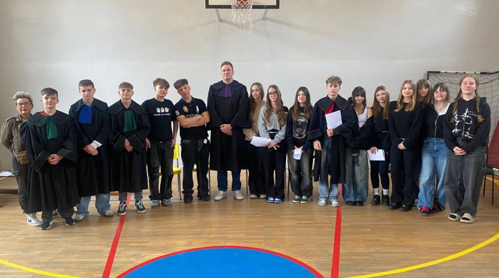 Uczniowie klas 7 i 8 wraz z nauczycielem pozują w sali gimnastycznej po zajęciach z edukacji prawnej, część z nich ubrana jest w togi i trzyma materiały związane z symulacją rozprawy sądowej.