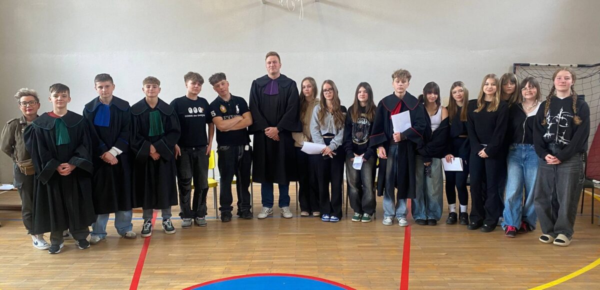 Uczniowie klas 7 i 8 wraz z nauczycielem pozują w sali gimnastycznej po zajęciach z edukacji prawnej, część z nich ubrana jest w togi i trzyma materiały związane z symulacją rozprawy sądowej.