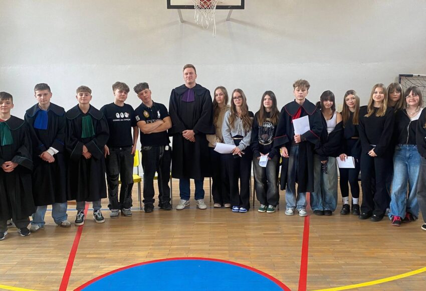 Uczniowie klas 7 i 8 wraz z nauczycielem pozują w sali gimnastycznej po zajęciach z edukacji prawnej, część z nich ubrana jest w togi i trzyma materiały związane z symulacją rozprawy sądowej.