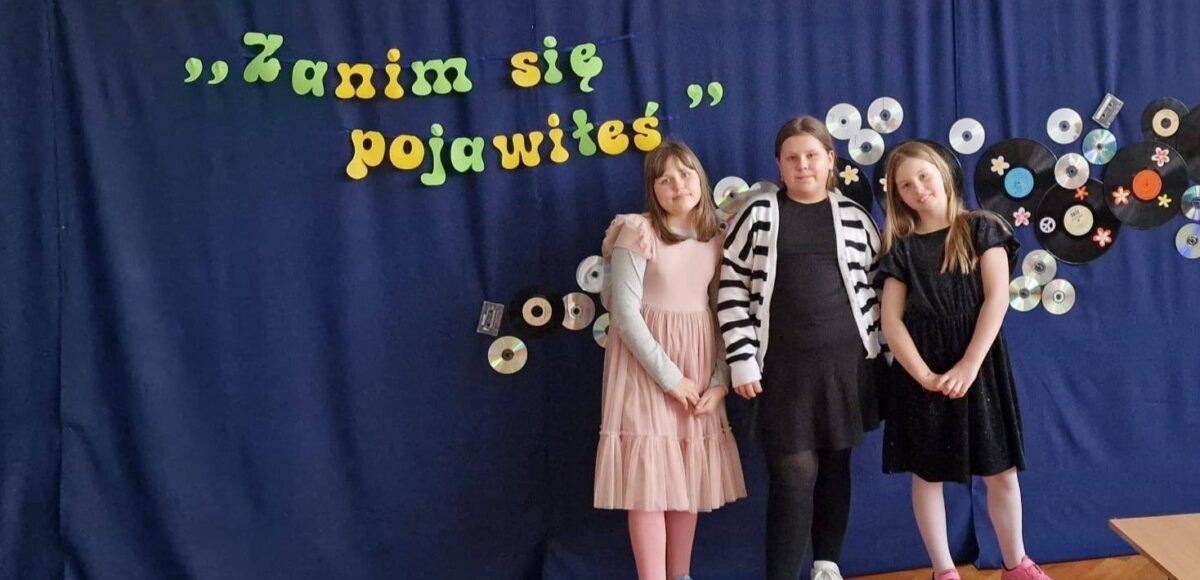 Uczennice szkoły podczas powiatowego Festiwalu Piosenki Polskiej „Zanim się pojawiłeś…”, pozujące na tle scenicznej dekoracji z napisem festiwalu.