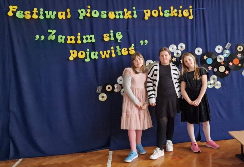 Uczennice szkoły podczas powiatowego Festiwalu Piosenki Polskiej „Zanim się pojawiłeś…”, pozujące na tle scenicznej dekoracji z napisem festiwalu.