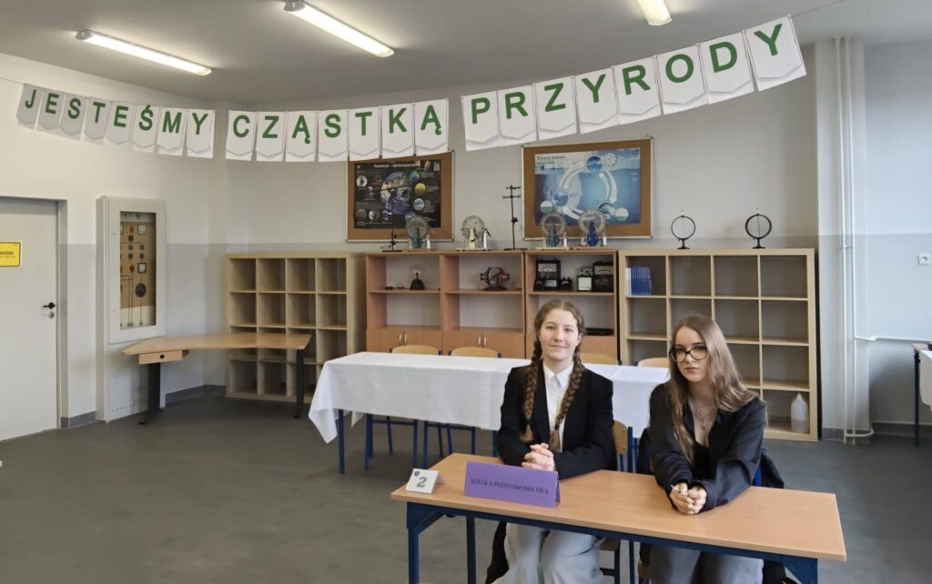 Uczennice klasy 8 reprezentujące szkołę podczas powiatowego konkursu „Jesteśmy cząstką przyrody”, siedzące przy stoliku konkursowym w sali przygotowanej do rywalizacji.