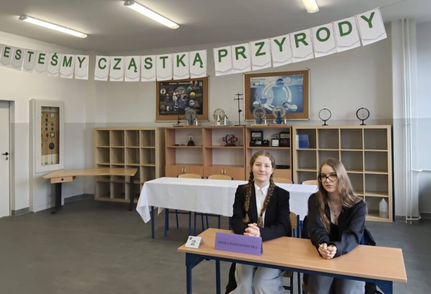 Uczennice klasy 8 reprezentujące szkołę podczas powiatowego konkursu „Jesteśmy cząstką przyrody”, siedzące przy stoliku konkursowym w sali przygotowanej do rywalizacji.