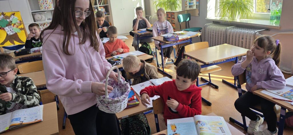 Uczniowie w klasie podczas obchodów Prima Aprilis losują „tajne kody” z koszyka, uczestnicząc w przygotowanej przez Samorząd Uczniowski zabawie.