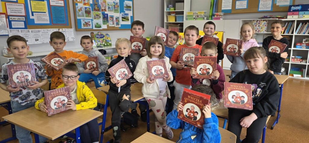 Uczniowie klasy pierwszej stoją w sali lekcyjnej i prezentują książki, które otrzymali po wizycie w bibliotece.