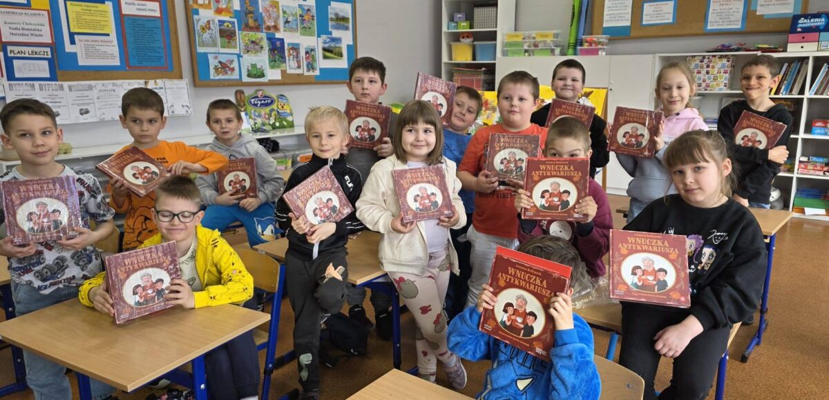 Uczniowie klasy pierwszej stoją w sali lekcyjnej i prezentują książki, które otrzymali po wizycie w bibliotece.