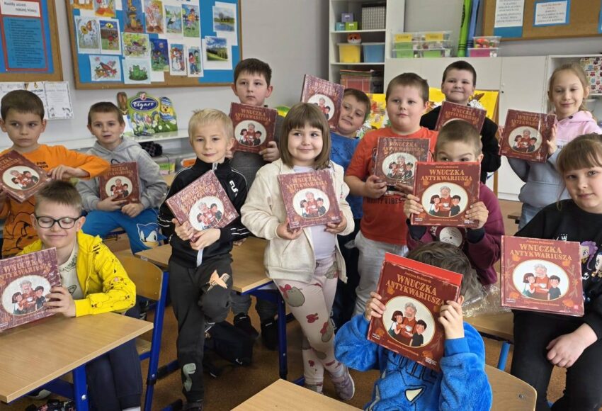 Uczniowie klasy pierwszej stoją w sali lekcyjnej i prezentują książki, które otrzymali po wizycie w bibliotece.