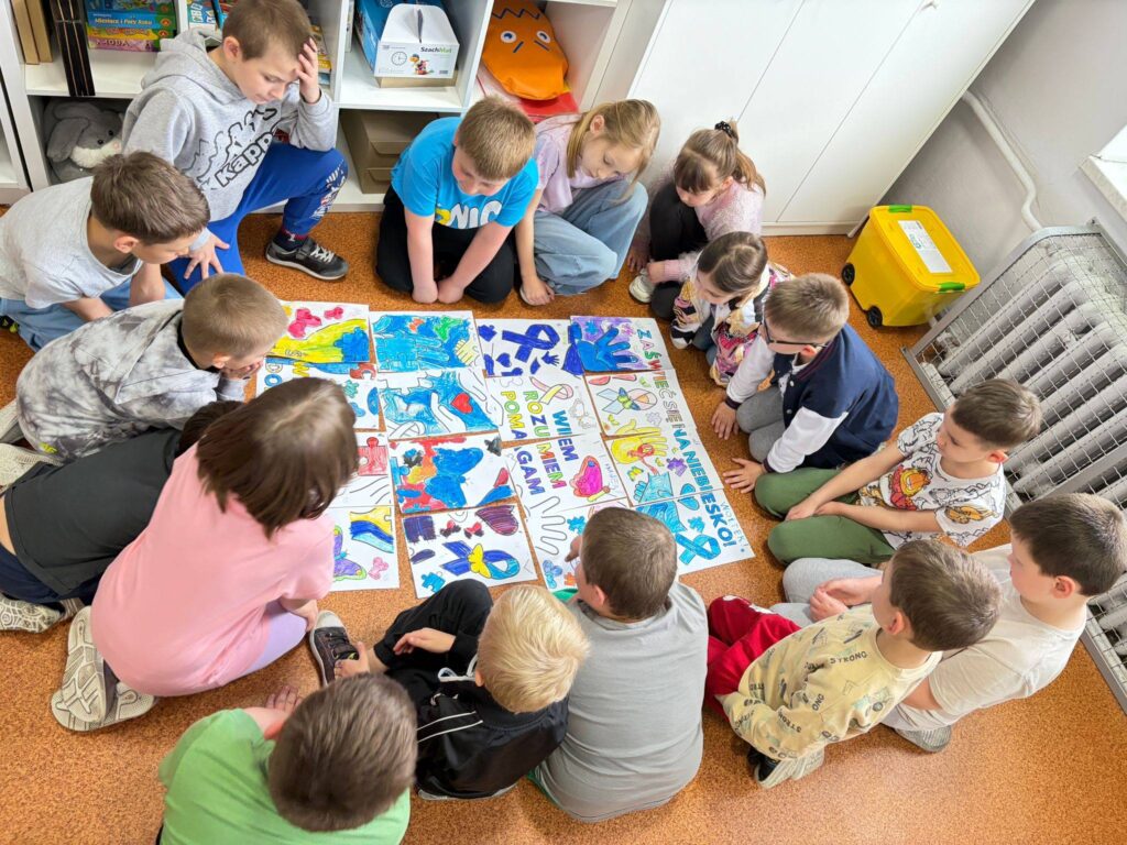 Uczniowie siedzą w kręgu na podłodze i wspólnie oglądają kolorowe prace plastyczne ułożone w formie puzzli, wykonane z okazji Światowego Dnia Świadomości Autyzmu.