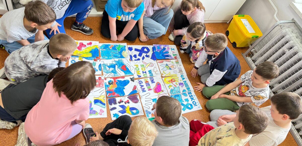Uczniowie siedzą w kręgu na podłodze i wspólnie oglądają kolorowe prace plastyczne ułożone w formie puzzli, wykonane z okazji Światowego Dnia Świadomości Autyzmu.