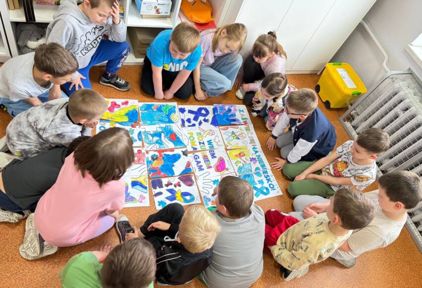 Uczniowie siedzą w kręgu na podłodze i wspólnie oglądają kolorowe prace plastyczne ułożone w formie puzzli, wykonane z okazji Światowego Dnia Świadomości Autyzmu.