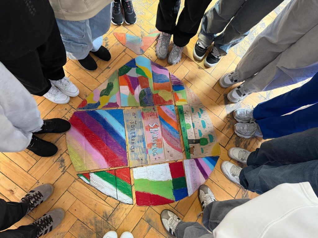 Uczniowie stoją w kręgu wokół wspólnej pracy plastycznej przedstawiającej kolorowe elementy symbolizujące różnorodność i współpracę.