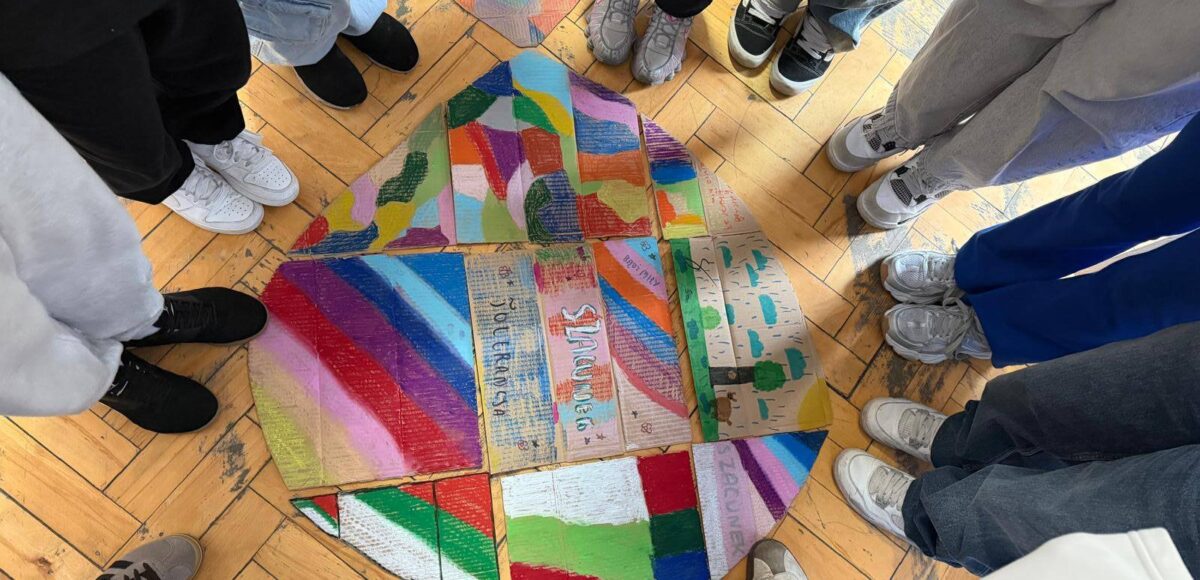 Uczniowie stoją w kręgu wokół wspólnej pracy plastycznej przedstawiającej kolorowe elementy symbolizujące różnorodność i współpracę.