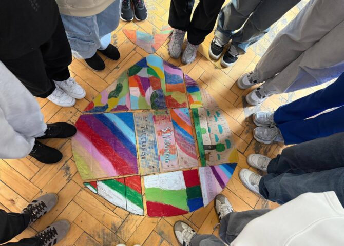 Uczniowie stoją w kręgu wokół wspólnej pracy plastycznej przedstawiającej kolorowe elementy symbolizujące różnorodność i współpracę.