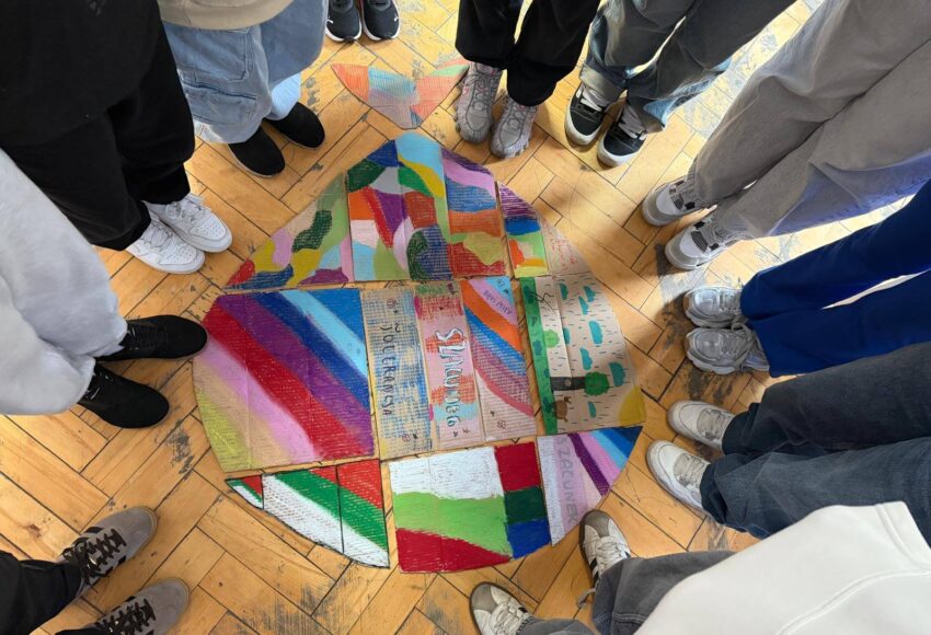 Uczniowie stoją w kręgu wokół wspólnej pracy plastycznej przedstawiającej kolorowe elementy symbolizujące różnorodność i współpracę.