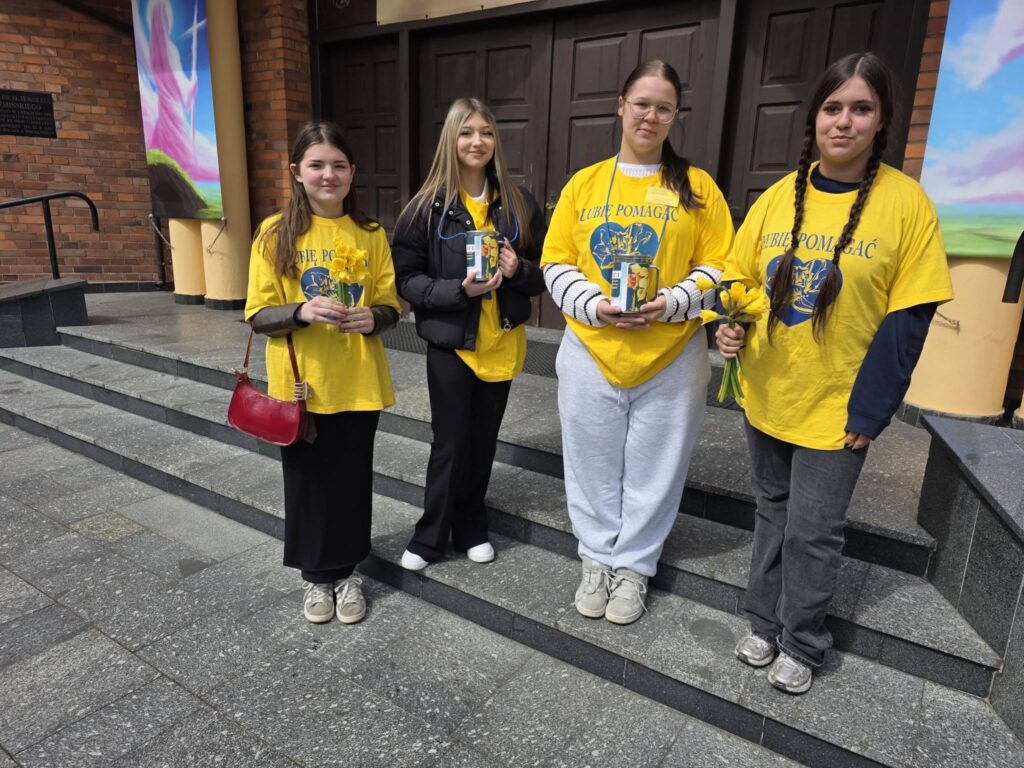 Wolontariusze ze szkoły stoją przed budynkiem i trzymają żonkile oraz puszki podczas akcji charytatywnej na rzecz hospicjum.