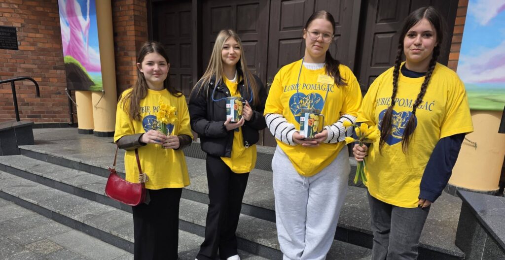 Wolontariusze ze szkoły stoją przed budynkiem i trzymają żonkile oraz puszki podczas akcji charytatywnej na rzecz hospicjum.