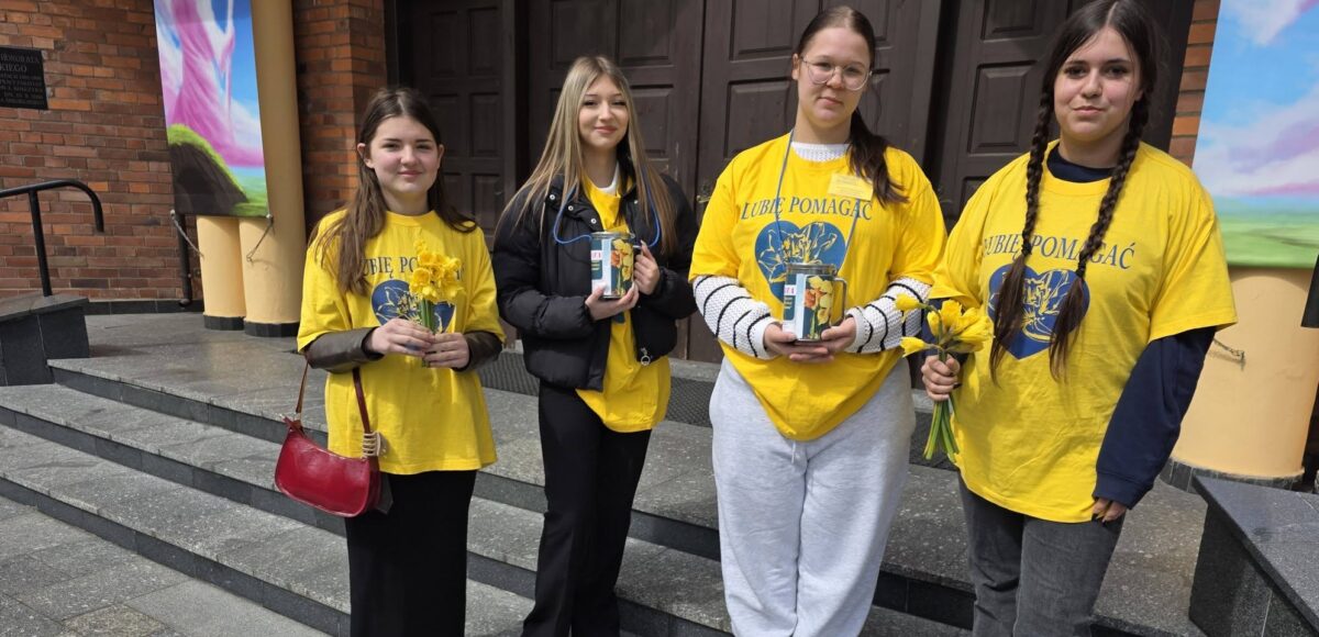 Wolontariusze ze szkoły stoją przed budynkiem i trzymają żonkile oraz puszki podczas akcji charytatywnej na rzecz hospicjum.