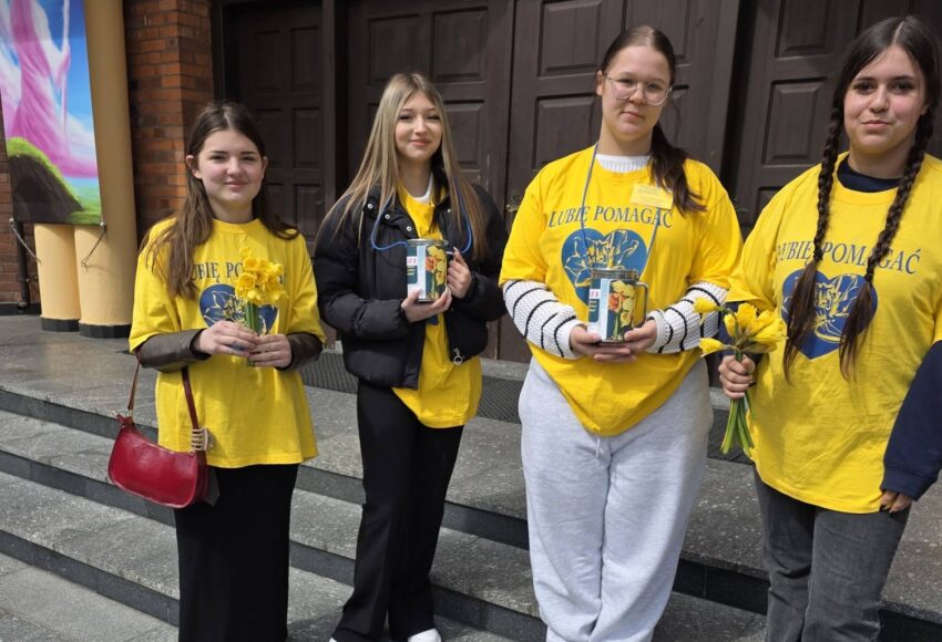 Wolontariusze ze szkoły stoją przed budynkiem i trzymają żonkile oraz puszki podczas akcji charytatywnej na rzecz hospicjum.