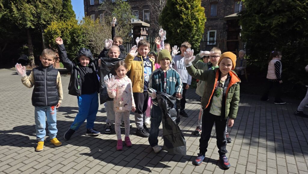 Uczniowie sprzątają teren wokół szkoły podczas obchodów Dnia Ziemi, trzymając worki i rękawice ochronne.