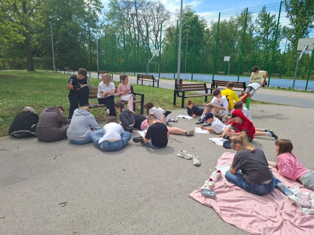 Uczniowie klasy IV a podczas zajęć outdoor learningu na terenie szkoły, pracujący w grupach na szkolnym boisku podczas lekcji języka polskiego i języka angielskiego.