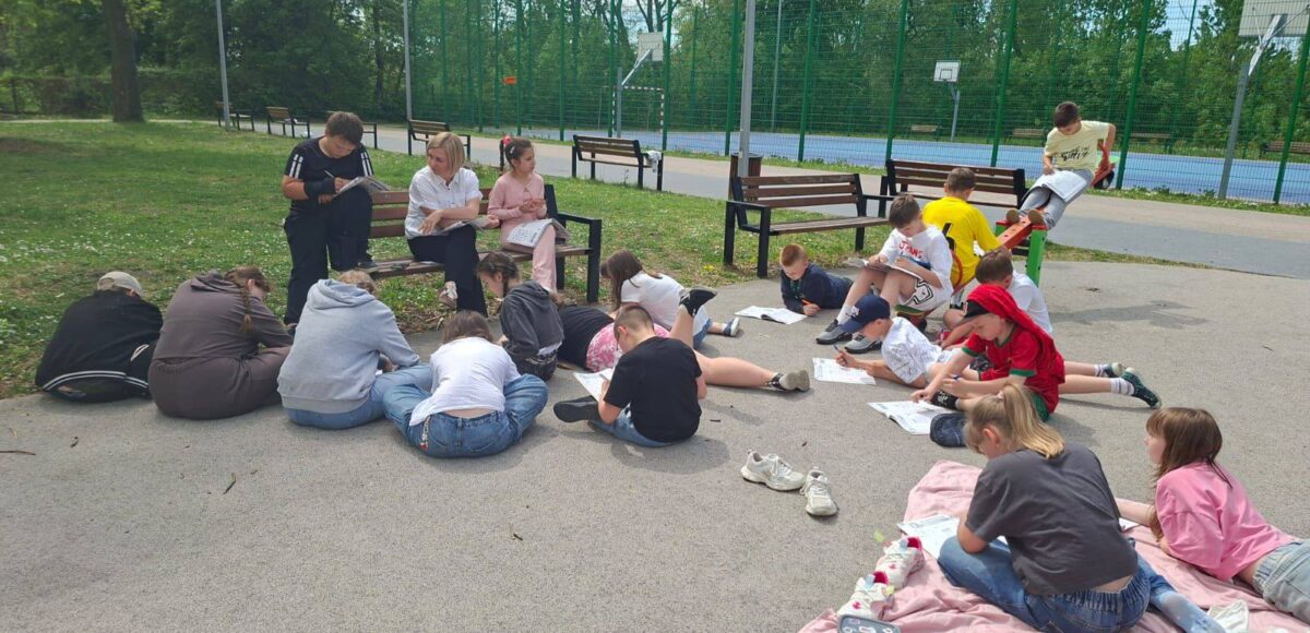 Uczniowie klasy IV a podczas zajęć outdoor learningu na terenie szkoły, pracujący w grupach na szkolnym boisku podczas lekcji języka polskiego i języka angielskiego.
