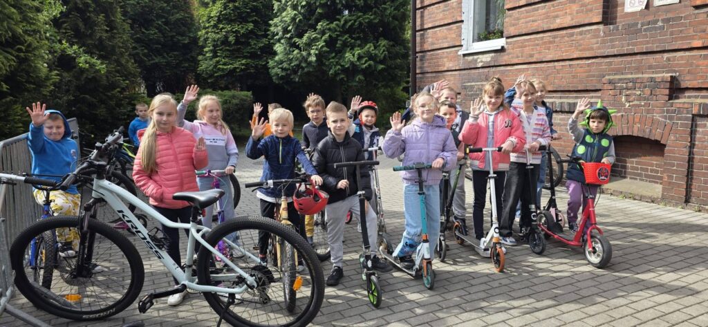 Uczniowie przed budynkiem szkoły uczestniczący w akcji „Kręci mnie maj”. Dzieci stoją z rowerami i hulajnogami, promując aktywny i ekologiczny dojazd do szkoły.