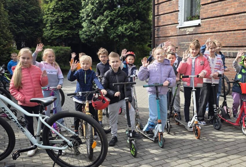 Uczniowie przed budynkiem szkoły uczestniczący w akcji „Kręci mnie maj”. Dzieci stoją z rowerami i hulajnogami, promując aktywny i ekologiczny dojazd do szkoły.