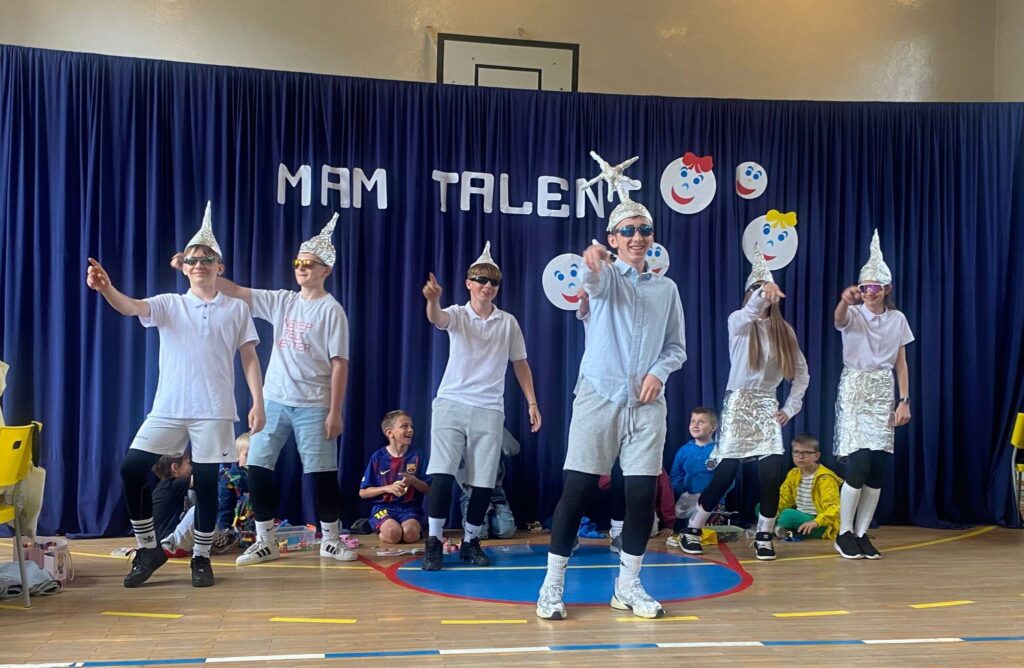 Uczniowie podczas występu tanecznego w czasie szkolnego wydarzenia „Mam Talent!”. Grupa prezentuje układ choreograficzny na sali gimnastycznej na tle dekoracji z napisem „Mam Talent”.