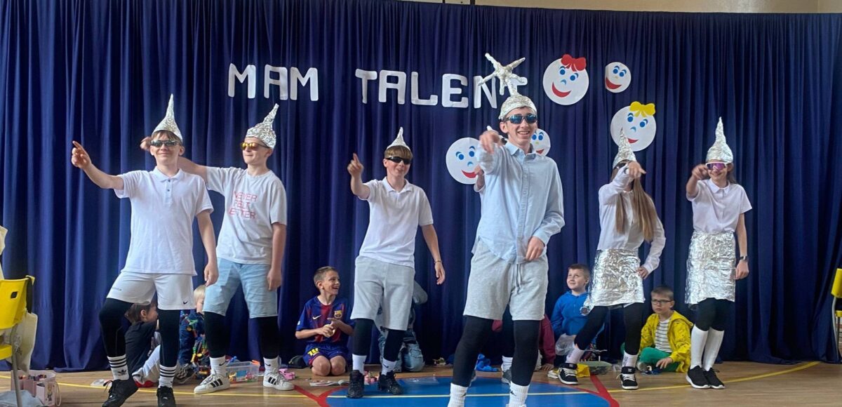Uczniowie podczas występu tanecznego w czasie szkolnego wydarzenia „Mam Talent!”. Grupa prezentuje układ choreograficzny na sali gimnastycznej na tle dekoracji z napisem „Mam Talent”.