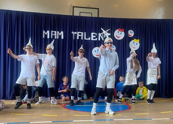 Uczniowie podczas występu tanecznego w czasie szkolnego wydarzenia „Mam Talent!”. Grupa prezentuje układ choreograficzny na sali gimnastycznej na tle dekoracji z napisem „Mam Talent”.