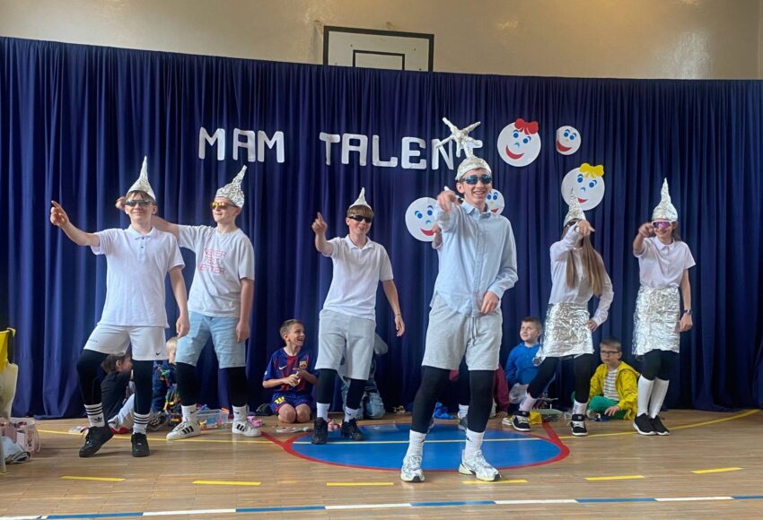 Uczniowie podczas występu tanecznego w czasie szkolnego wydarzenia „Mam Talent!”. Grupa prezentuje układ choreograficzny na sali gimnastycznej na tle dekoracji z napisem „Mam Talent”.