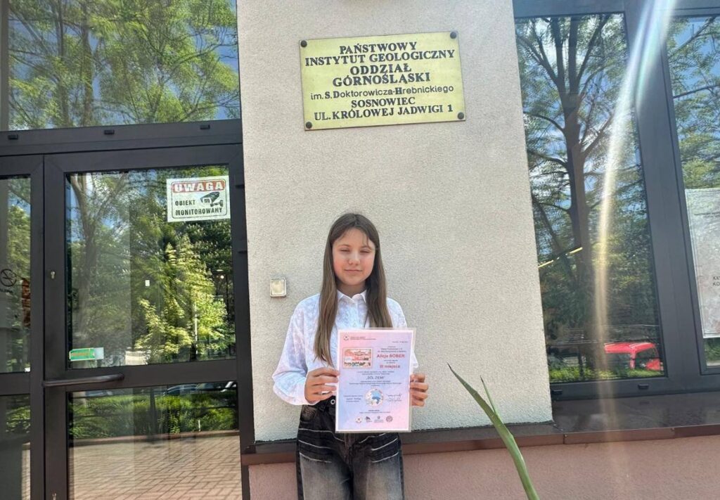 Uczennica klasy VI prezentuje dyplom za zdobycie III miejsca w półfinale regionalnym Ogólnopolskiego Konkursu Plastycznego „Sól Ziemi” przed budynkiem Państwowego Instytutu Geologicznego – Oddziału Górnośląskiego.